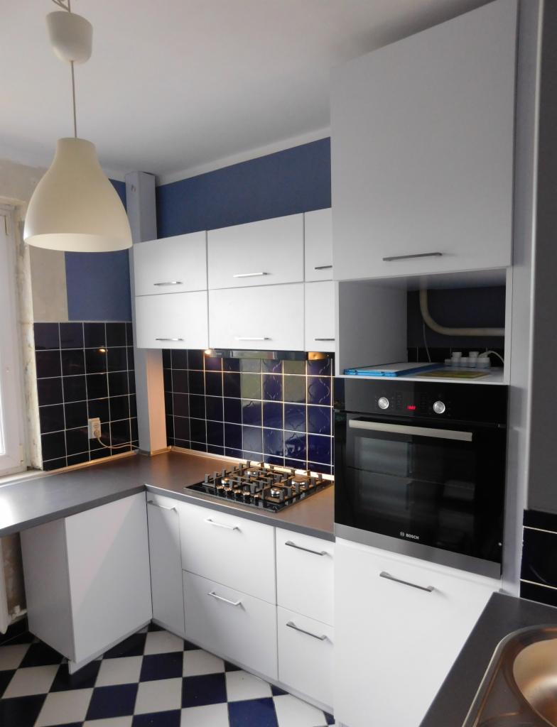 Biała kuchnia z niebieskimi akcentami: szafki wiszące i stojące, piekarnik Bosch, płyta gazowa. Czarno-niebieska mozaika na ścianie i podłodze.