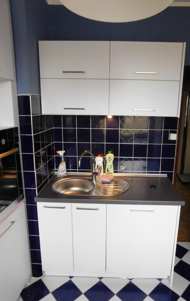 Biała szafka pod zlew z ciemnoszarym blatem i baterią, nad nią wiszące szafki. Na blacie płyny do naczyń. Tło stanowią ciemnoniebieskie płytki i podłoga w biało-niebieską szachownicę.