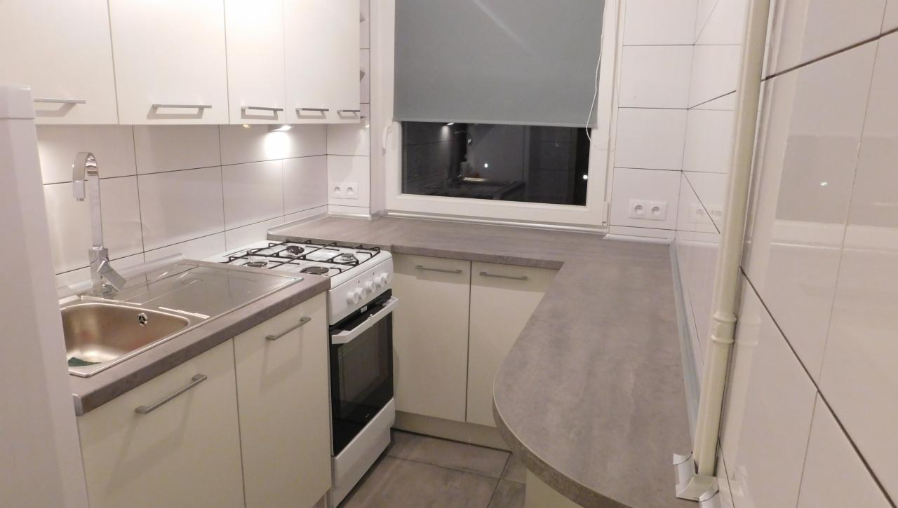 Jasna kuchnia z białymi szafkami, szarym blatem i płytkami. Widoczny zlew, kuchenka gazowa i okno z roletą. Minimalistyczny design, funkcjonalne rozwiązanie.