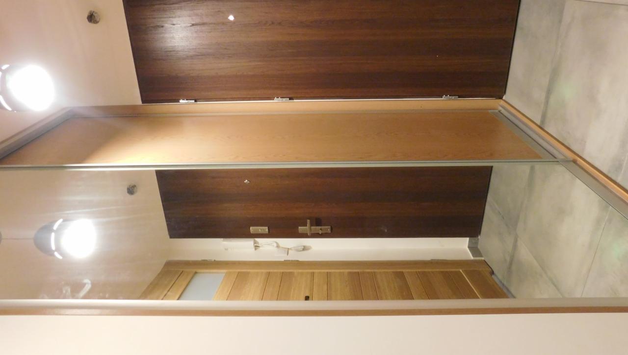 Jasne drewno szafy przesuwnej z lustrem odbija fragment pokoju: drzwi z szybą, sufit z lampami i ciemne drewno. Widoczne metalowe okucia i szara ściana.