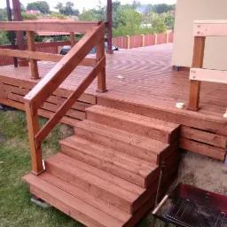 Mężczyzna maluje drewnianą konstrukcję tarasu z schodami, widoczny grill obok schodów, w tle dom jednorodzinny i ogrodzenie.