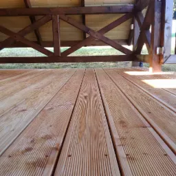 Świeżo położona, jasna deska podłogowa z drewna na zewnątrz, widok z niskiej perspektywy, ukazujący strukturę drewna i elementy konstrukcyjne z belek w tle.