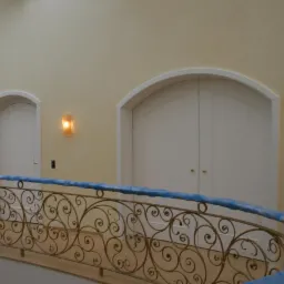 Białe, dwuskrzydłowe drzwi z łukiem, oświetlone kinkietami, widoczne zza metalowej, zdobionej balustrady na tle kremowej ściany w eleganckim wnętrzu.