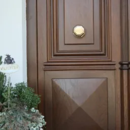 Ciemnobrązowe, drewniane drzwi wejściowe z dekoracyjnym okuciem i napisem 'Välkommen' obok donicy z roślinami, przy białej ścianie.