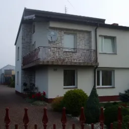 Dwupiętrowy dom z balkonem i numerem 29, otynkowany na biało z elementami kamiennej elewacji, antena satelitarna na ścianie, zadbany ogród z przodu.