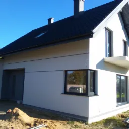 Nowoczesny dom jednorodzinny z jasną elewacją, ciemną dachówką i szarymi oknami, widok z zewnątrz w słoneczny dzień, z widoczną stertą piasku przy ścianie.