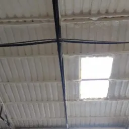 Wnętrze hali przemysłowej z metalową konstrukcją dachu pokrytego grubą warstwą białej pianki izolacyjnej, widoczne fragmenty oświetlenia i okno dachowe.