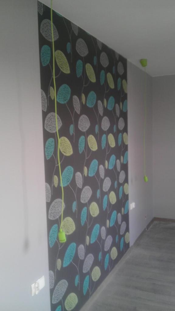 Ściana z tapetą w pastelowe liście, zielony kabel zwisający z sufitu. Ukończone wykończenie wnętrza, widoczne gniazdko elektryczne.