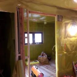 Wnętrze w trakcie ocieplania, ściany i sufit pokryte żółtą folią izolacyjną, widoczne drewniane ramy konstrukcyjne, okno, nagrzewnica elektryczna i porozrzucane przewody.