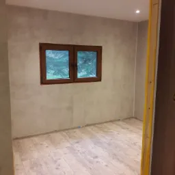 Pok&oacute;j po remoncie z nową podłogą laminowaną, świeżo pomalowanymi ścianami w odcieniach szarości i drewnianym oknem z widokiem na zieleń, widoczne listwy przypodłogowe.