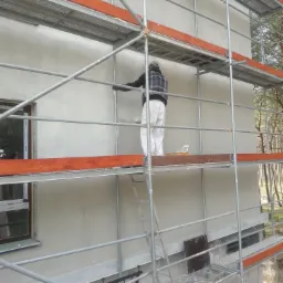 Pracownik w odzieży ochronnej na rusztowaniu maluje elewację budynku wałkiem; widoczny fragment okna i las w tle.