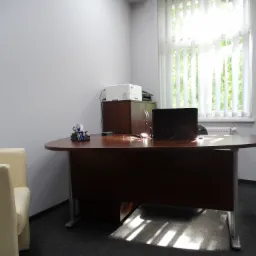 Jasne biuro z ciemnobrązowym biurkiem, krzesłem w kolorze ecru i oknem z pionowymi żaluzjami, drukarka na szafce w tle.