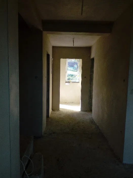 Korytarz w surowym stanie z otynkowanymi ścianami, widoczne otwory drzwiowe i okno w głębi, betonowa podłoga z resztkami materiałów budowlanych, surowe oświetlenie.