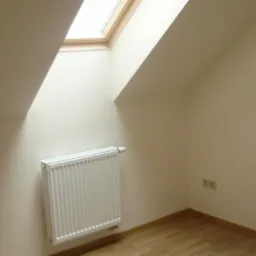 Poddasze z oknem dachowym, grzejnikiem panelowym i parkietem ułożonym w jodełkę, ściany w odcieniach beżu i widoczne gniazdka elektryczne.