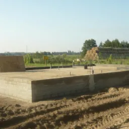 Wylane fundamenty pod budynek jednorodzinny z widocznymi zbrojeniami i wyprowadzonymi rurami, w tle stóg siana i zabudowania gospodarcze w Rożnowie.