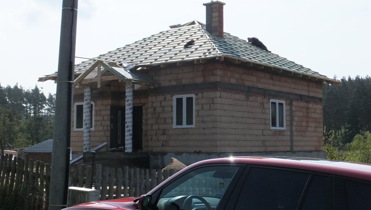 Budowa domu murowanego w trakcie realizacji: widoczne ściany z pustaków, okna PCV z folią ochronną, więźba dachowa i komin w stanie surowym, z przodu częściowo zasłonięte przez czerwony samochód.