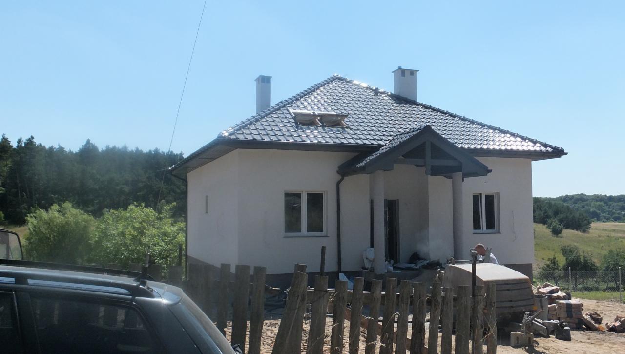 Niedokończony dom jednorodzinny z szarą dachówką, widoczne materiały budowlane i częściowo postawione ogrodzenie z drewnianych sztachet.