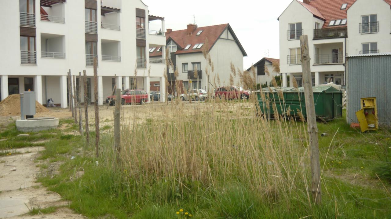 Teren budowy z widocznymi budynkami mieszkalnymi w tle, ogrodzony drewnianym płotem z drutem kolczastym, zarośnięty trawą i chwastami, z kontenerem i skrzynką elektryczną, w trakcie przygotowania...