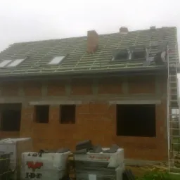 Budowa domu w trakcie: widoczne krokwie, okna dachowe, komin i częściowo ułożona dachówka. Na dachu pracownik, przy budynku drabina i palety z materiałami.