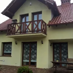 Dwupiętrowy dom z balkonem i czerwoną dachówką, widok z zewnątrz. Rynny w kolorze dachu. Obok budynek z panelami słonecznymi na dachu.