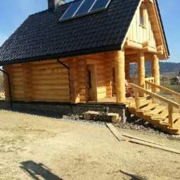 Wykończony dom z bali z czarnym dachem i panelami słonecznymi, widoczny z zewnątrz na tle pagórkowatego krajobrazu.