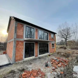 Surowy stan budynku w trakcie budowy z zamontowanymi oknami z czarnymi ramami. W tle drzewa bez liści i fragmenty innych budynków.