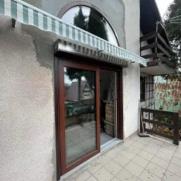 Brązowe okno przesuwne PCV z zamontowaną roletą zewnętrzną w kolorze biało-zielonym na elewacji domu w trakcie wykończenia, widoczny balkon z drewnianą balustradą oraz stos drewna.