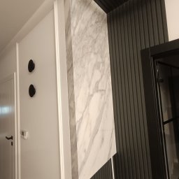 ERBUD ERNEST ŻABICKI - Nowoczesna aranżacja wnętrza: biała ściana z czarnymi wieszakami, marmurowa kolumna i ciemna, żłobkowana ściana. Minimalistyczny design z wyraźnymi kontrastami kolorystycznymi.