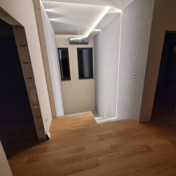 ERBUD ERNEST ŻABICKI - Nowoczesne wykończenie wnętrza: drewniana podłoga, ściany w jasnych odcieniach z subtelnym oświetleniem LED i klimatyzacja. Widoczne fragmenty drzwi i okien.