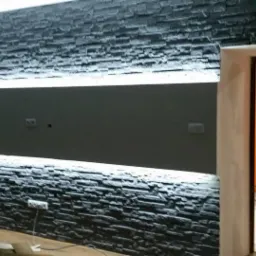 Dekoracyjna ściana z czarnej okładziny kamiennej z poziomym pasem podświetlenia LED, widok na pomarańczowy korytarz przez otwarte drzwi.