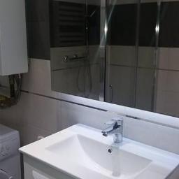 ERBUD ERNEST ŻABICKI - Nowoczesna łazienka z białą umywalką nablatową, szafką z frezowanymi uchwytami, kabiną prysznicową ze srebrnymi profilami i pralką z boku, oświetlona listwą LED pod lustrem.