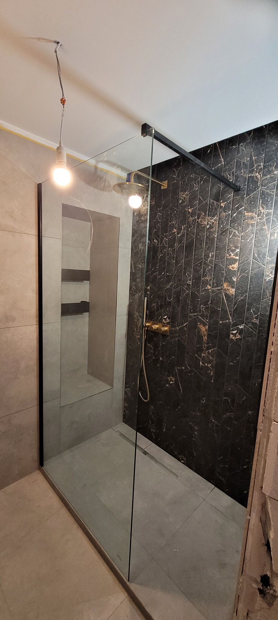 Nowoczesna kabina prysznicowa z czarnymi profilami i deszczownicą w złotej armaturze, na tle ściany z ciemnych płytek imitujących marmur. Widoczna wnęka z półkami.