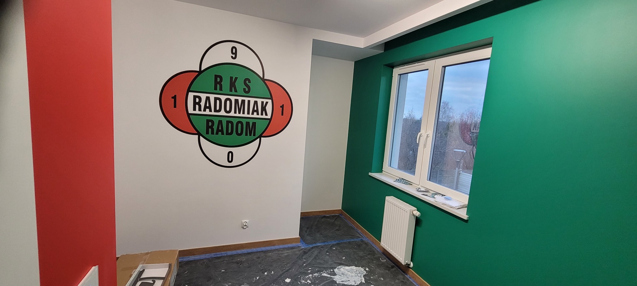Wnętrze po malowaniu: ściana z logo klubu sportowego RKS Radomiak Radom, obok okno i grzejnik. Ściany w kolorach biały, czerwony i zielony. Podłoga zabezpieczona folią.