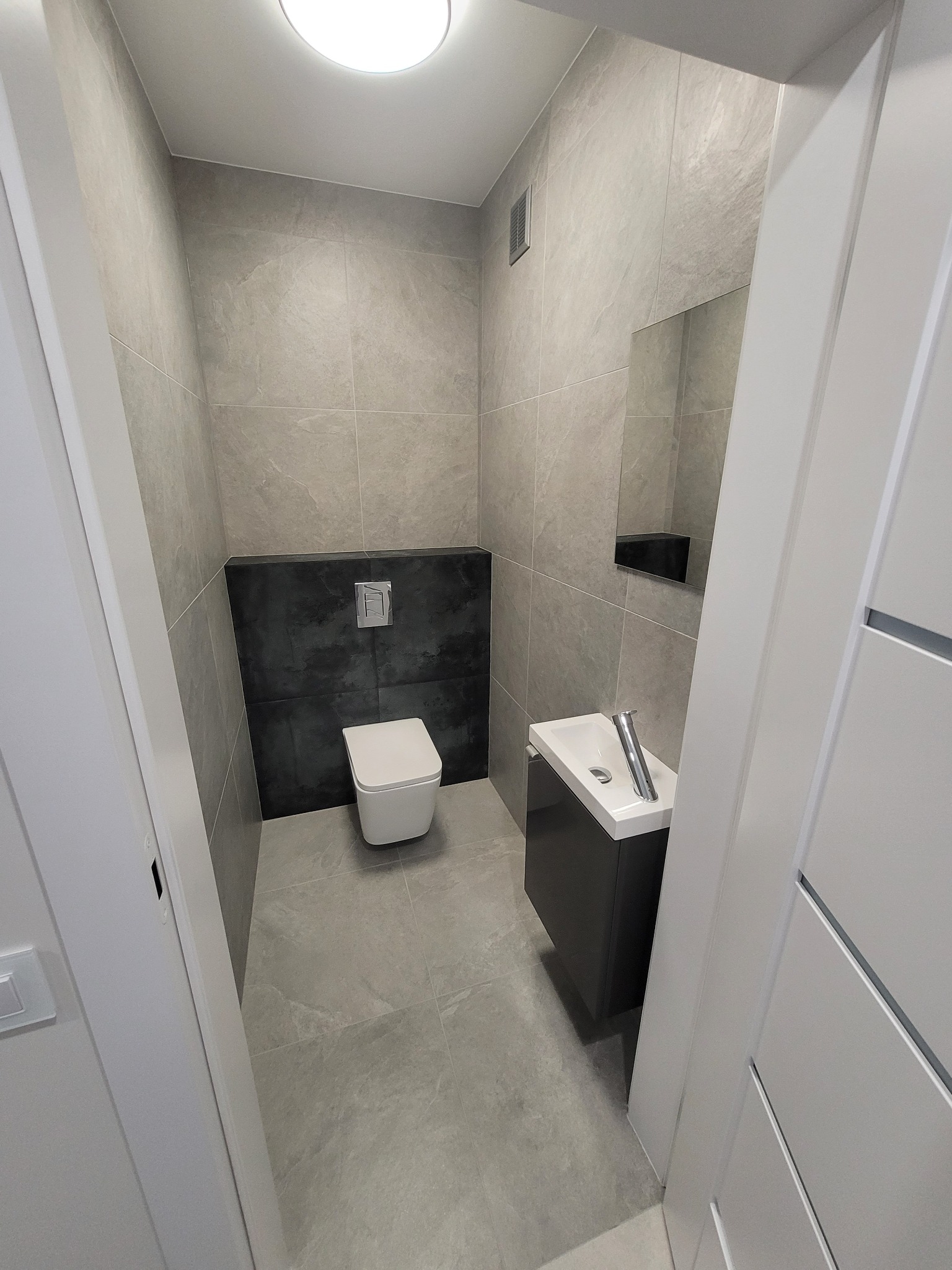 Nowoczesna łazienka w szarościach z podwieszaną toaletą i umywalką na szafce. Minimalistyczny design, płytki imitujące kamień, czarny akcent za toaletą.