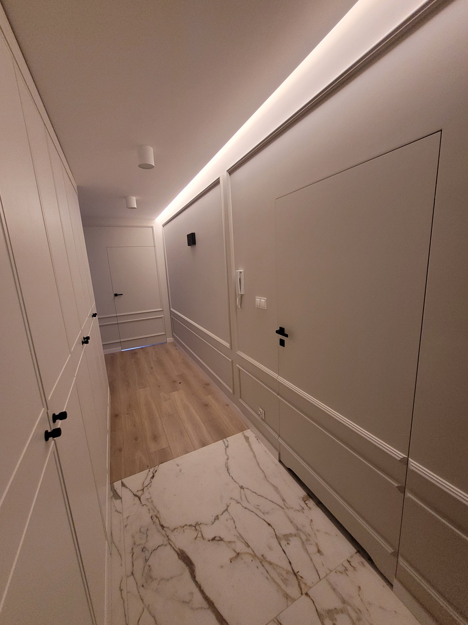 Elegancki korytarz po remoncie: białe ściany z dekoracyjnymi panelami, ukryte drzwi z czarnymi klamkami, marmurowa podłoga i oświetlenie LED. Nowoczesny design.