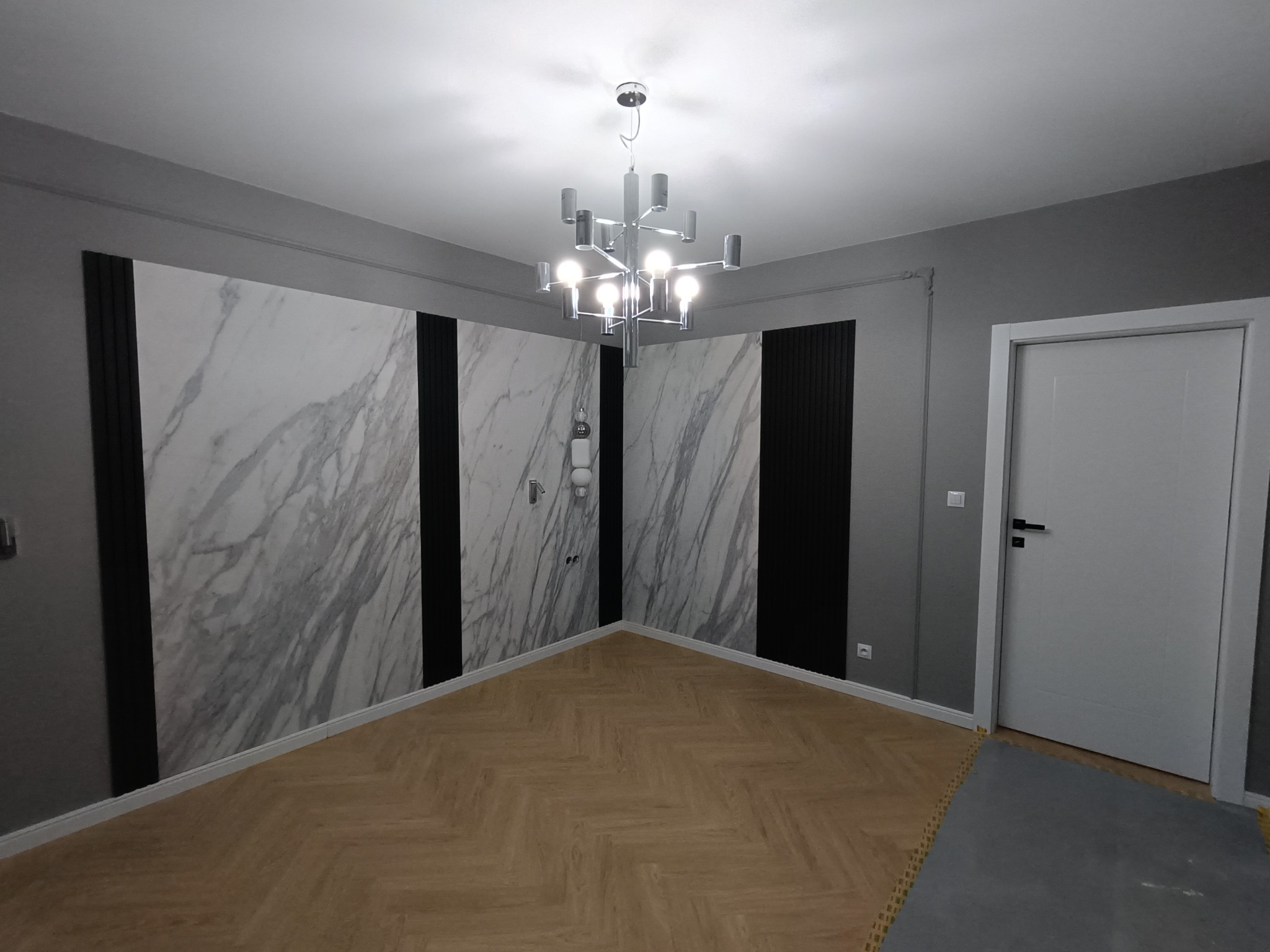 Nowoczesne wnętrze: parkiet w jodełkę, ściany z marmurowymi panelami i czarnymi wstawkami, designerski żyrandol. Minimalistyczny styl, szaro-biała kolorystyka.