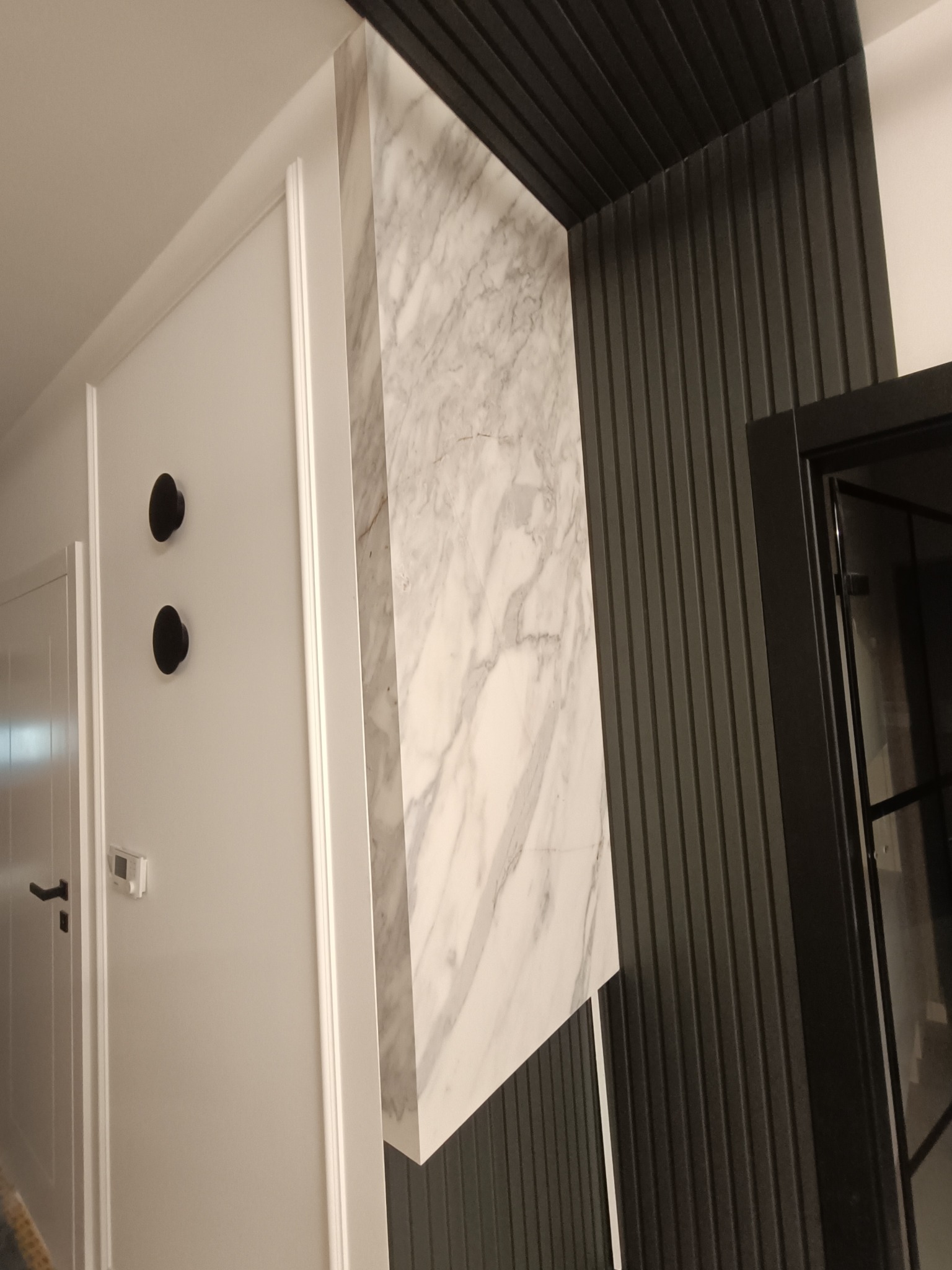 Nowoczesna aranżacja wnętrza: biała ściana z czarnymi wieszakami, marmurowa kolumna i ciemna, żłobkowana ściana. Minimalistyczny design z wyraźnymi kontrastami kolorystycznymi.