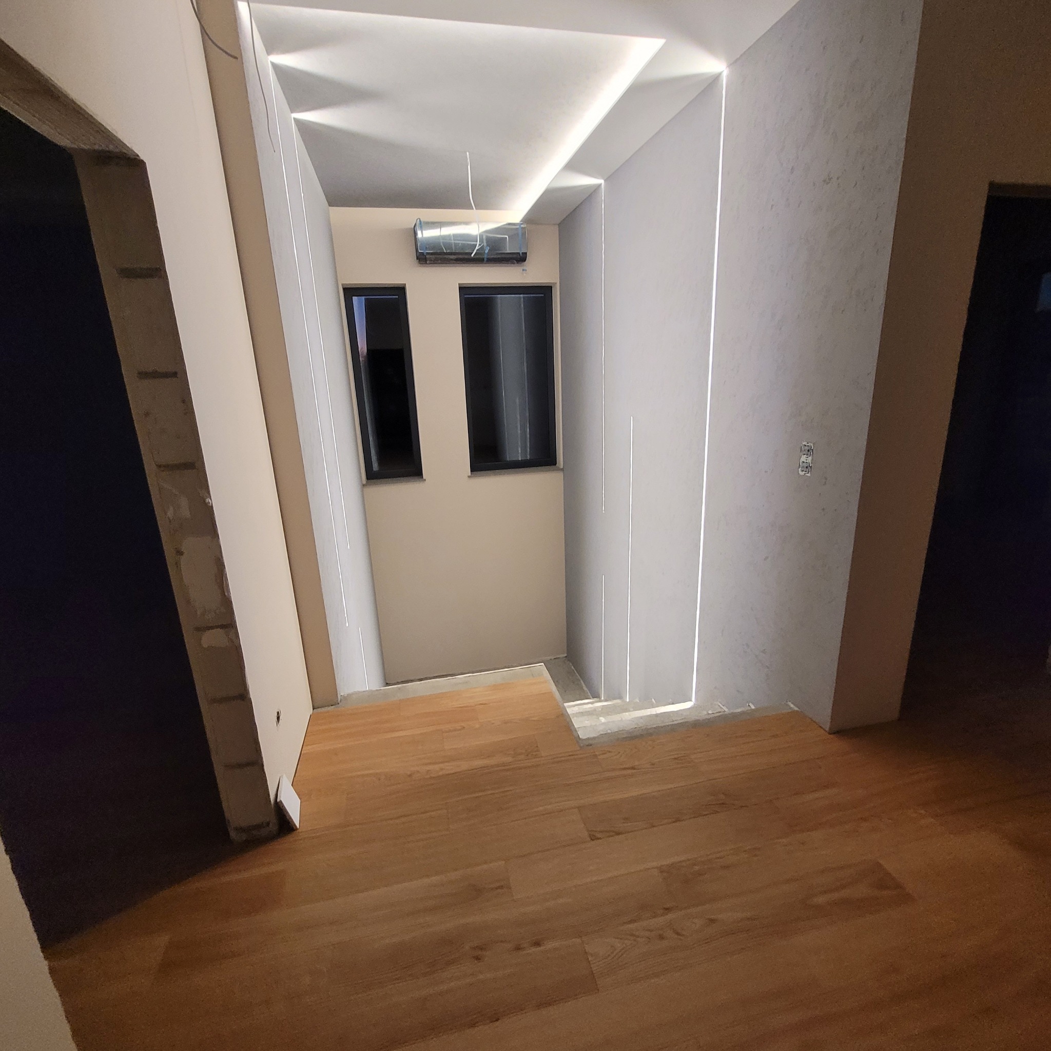 Nowoczesne wykończenie wnętrza: drewniana podłoga, ściany w jasnych odcieniach z subtelnym oświetleniem LED i klimatyzacja. Widoczne fragmenty drzwi i okien.