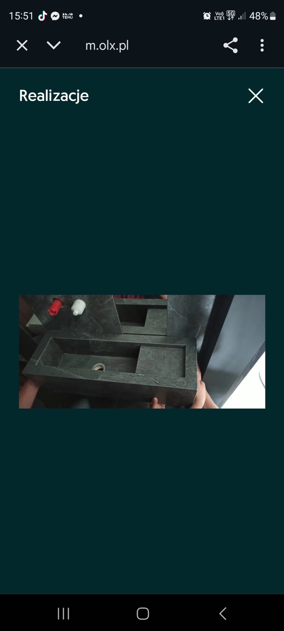 Wyjątkowa, kamienna umywalka w odcieniach szarości, z widocznymi elementami instalacji wodnej. Umywalka o geometrycznym kształcie, z wyraźną fakturą kamienia.