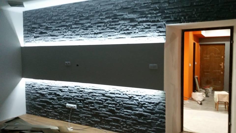 Dekoracyjna ściana z czarnej okładziny kamiennej z poziomym pasem podświetlenia LED, widok na pomarańczowy korytarz przez otwarte drzwi.