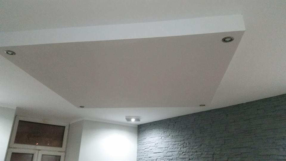 Podwieszany sufit z płyt gipsowo-kartonowych z czterema punktowymi oprawami oświetleniowymi, widok z dołu na szarą ścianę z imitacji kamienia i okno.