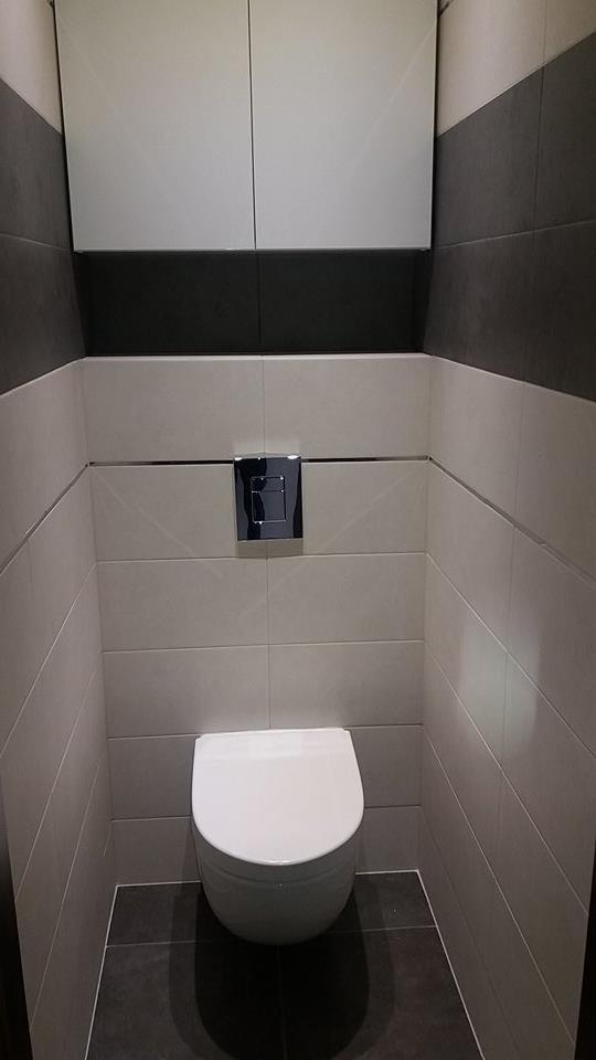 Nowoczesna toaleta podwieszana z chromowanym przyciskiem spłukującym, obudowana w szaro-białe płytki z ciemną podłogą i szafkami wiszącymi.