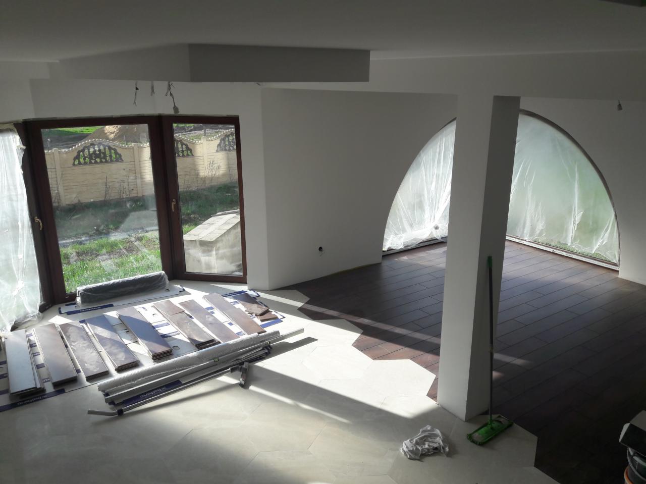 Układanie paneli podłogowych w jasnym, przestronnym wnętrzu z dużymi oknami, widoczne rozłożone panele i narzędzia, częściowo ułożona podłoga z ciemnego drewna, okna zabezpieczone folią ochronną.