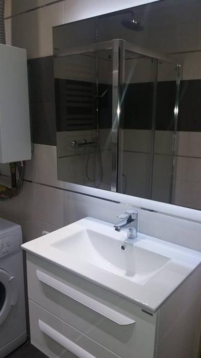 Nowoczesna łazienka z białą umywalką nablatową, szafką z frezowanymi uchwytami, kabiną prysznicową ze srebrnymi profilami i pralką z boku, oświetlona listwą LED pod lustrem.