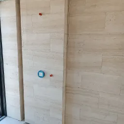 Ściana wyłożona beżowymi płytkami trawertynowymi, widoczne fragmenty instalacji ogrzewania podłogowego i wystające elementy instalacji wodnej, w trakcie prac wykończeniowych.