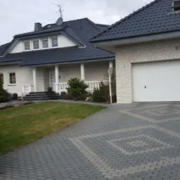 Elegancki dom jednorodzinny z garażem i podjazdem z kostki brukowej w Opolu. Jasna elewacja z ciemnym dachem i kolumnami przy wejściu.