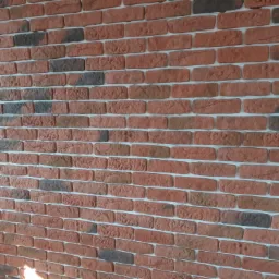 Dekoracyjna ściana z cegieł klinkierowych w odcieniach czerwieni i brązu, ułożonych z przesunięciem, z widocznymi fugami w jasnym kolorze.