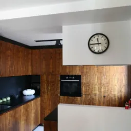 Nowoczesna kuchnia z drewnianymi szafkami o wyraźnym usłojeniu, czarnym blatem i elementami w kolorze czarnym i białym. Na ścianie zegar.