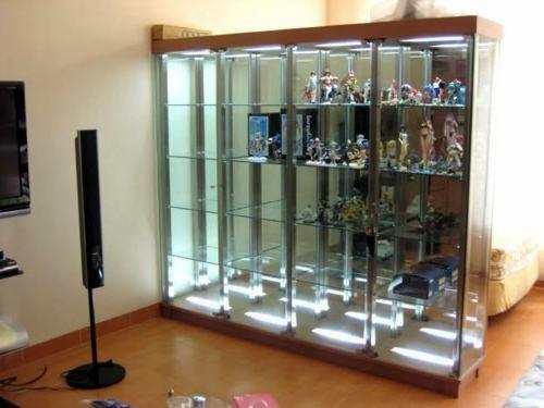 Szklana witryna ekspozycyjna z drewnianą ramą, oświetlona od góry, wypełniona kolekcją figurek i modeli. Widoczny fragment salonu z podłogą i sprzętem audio.