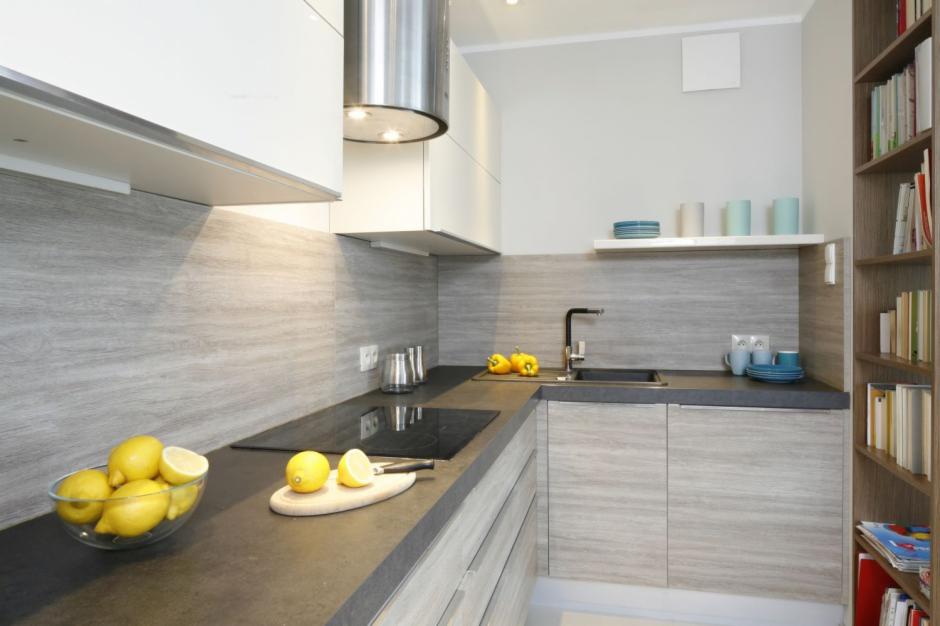 Nowoczesna kuchnia z szarymi płytkami imitującymi drewno na ścianie, czarnym blatem i białymi szafkami. Na blacie cytryny w misce i na desce. Minimalistyczny design.